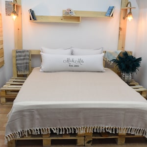 May include: A beige throw blanket with fringe edges is draped over a bed with a white pillow with the text "Mr. & Mrs. Est. 2015" on it. The bed is made of wooden pallets and is in a room with white walls and wooden shelves.