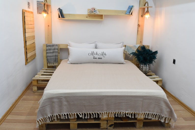 May include: A beige throw blanket with fringe edges is draped over a bed with a white pillow with the text "Mr. & Mrs. Est. 2015" on it. The bed is made of wooden pallets and is in a room with white walls and wooden shelves.