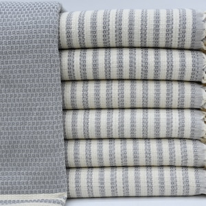 Puede incluir: Una pila de toallas rayadas grises y blancas con flecos. Las toallas están hechas de un material suave y absorbente y son perfectas para la playa o la piscina.
