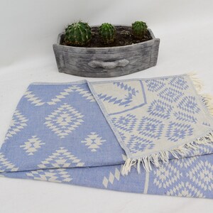 Peut inclure: Une serviette turque &agrave; franges avec un motif bleu clair et blanc. La serviette est pli&eacute;e en deux et pr&eacute;sente un motif g&eacute;om&eacute;trique.
