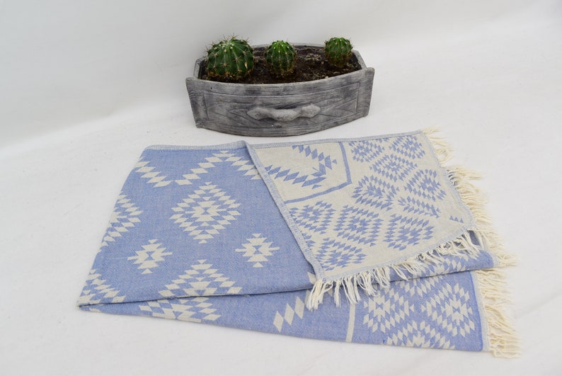 Peut inclure: Une serviette turque &agrave; franges avec un motif bleu clair et blanc. La serviette est pli&eacute;e en deux et pr&eacute;sente un motif g&eacute;om&eacute;trique.
