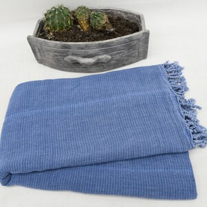 May include: A blue cotton throw blanket with fringe on the edges. The blanket is folded in half and lying on a white surface.