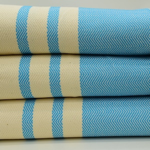 May include: A stack of four blue and white striped towels with fringe edges. The towels are made of cotton and have a herringbone pattern.