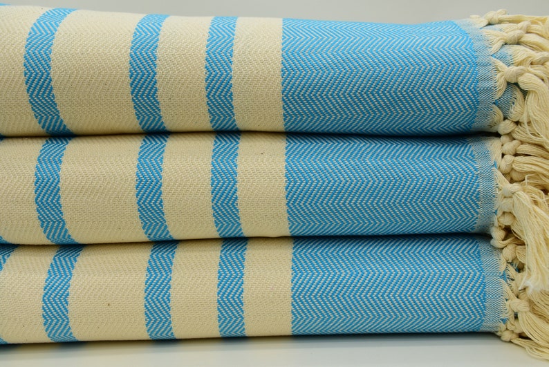 May include: A stack of four blue and white striped towels with fringe edges. The towels are made of cotton and have a herringbone pattern.