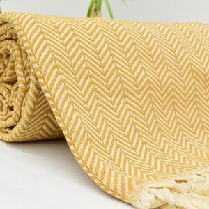 Op de afbeelding: Een gele en witte chevron-gepatroneerde deken met franjes. De deken is opgerold en heeft een zacht, textuur.