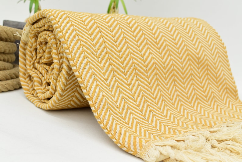 Op de afbeelding: Een gele en witte chevron-gepatroneerde deken met franjes. De deken is opgerold en heeft een zacht, textuur.