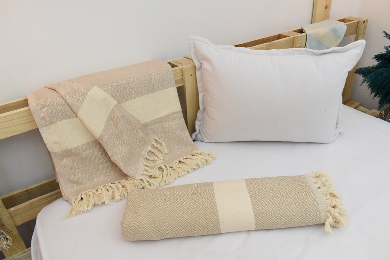 May include: Two beige and white striped throw blankets with fringe edges. One blanket is folded and the other is rolled up. The blankets are on a white bed with a white pillow.