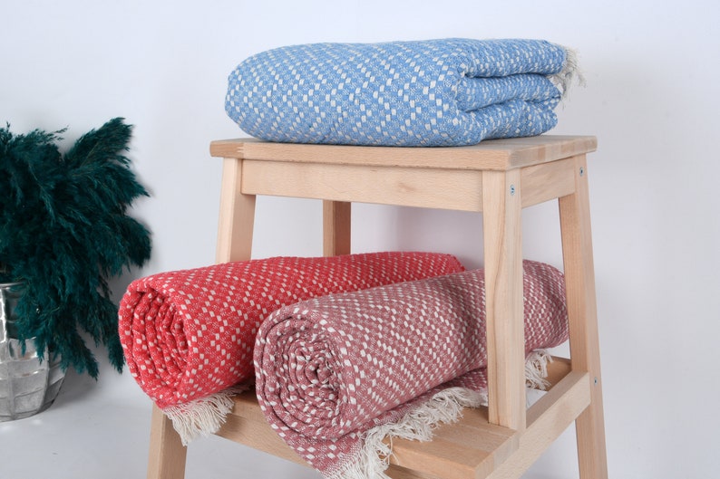 May include: Two rolled up blankets on a wooden stool. One blanket is a light blue with a white diamond pattern, and the other is a coral color with a white diamond pattern.