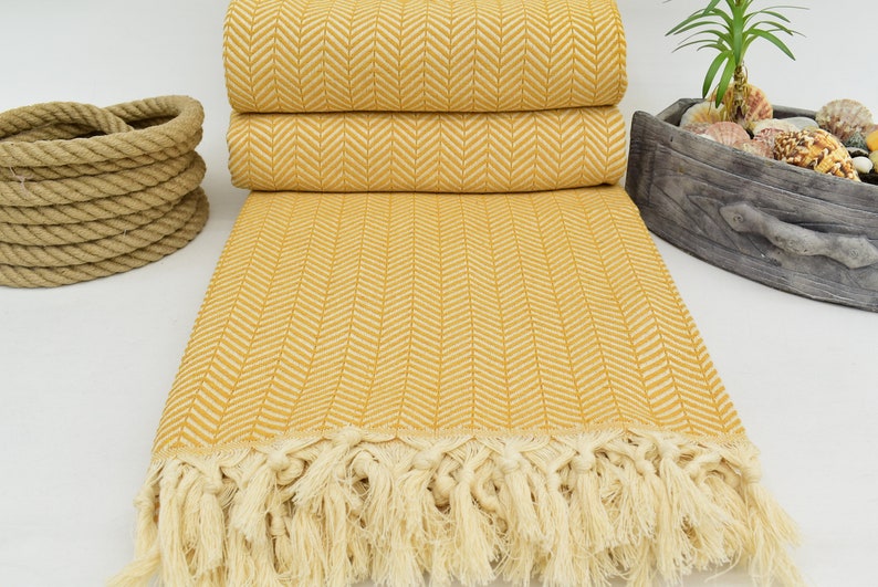Op de afbeelding: Een gele deken met een herringbone patroon en witte franjes. De deken is in drie&euml;n gevouwen en ligt op een wit oppervlak.