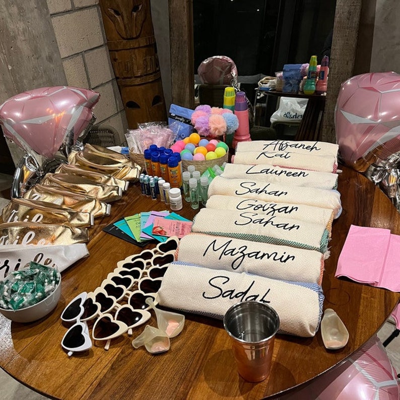 May include: A table laid out with various items for a bridal shower party. There are towels with names written on them, sunglasses, a bowl of sweets, and a pink balloon.