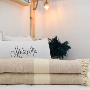 May include: Two beige and white striped throw blankets with fringe edges are folded on a white bed. The top blanket has a white pillow with the text "Mr. & Mrs." in black cursive writing behind it.
