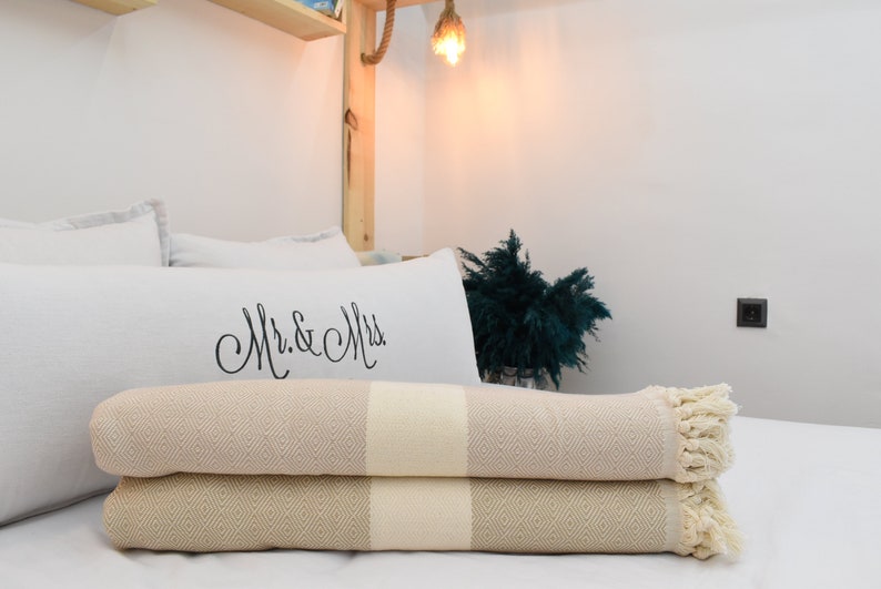 May include: Two beige and white striped throw blankets with fringe edges are folded on a white bed. The top blanket has a white pillow with the text "Mr. & Mrs." in black cursive writing behind it.