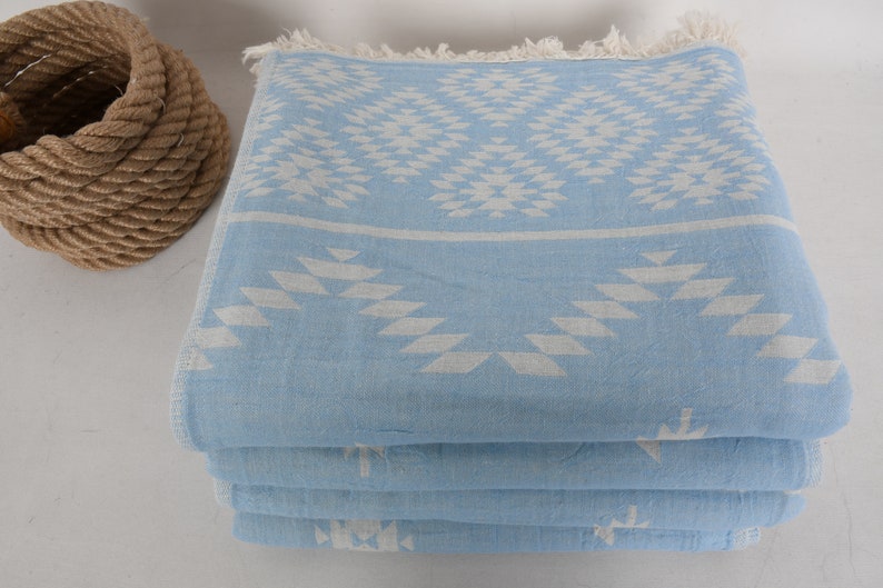 Pu&ograve; includere: Un mucchio di asciugamani di colore azzurro chiaro con un motivo geometrico bianco. Gli asciugamani sono piegati e hanno un bordo con frange.