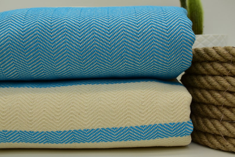 May include: Two folded Turkish towels with a chevron pattern. The top towel is turquoise blue and white, and the bottom towel is white with a turquoise blue border.