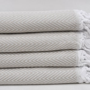 May include: A stack of four folded beige and white herringbone-patterned towels. The towels have a soft, textured appearance with white fringe at the ends. The neutral color palette suggests a spa or bathroom theme.
