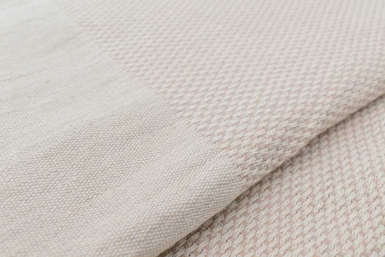May include: A close-up of a beige woven fabric with a subtle pattern. The fabric is soft and textured, with a slightly rough feel.