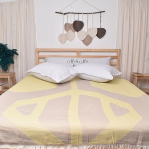 May include: A beige bed with a yellow and beige patterned duvet cover. Two white pillows with the text "Mr. & Mrs." are on the bed. The bed is in a bedroom with white curtains and a wall hanging with brown and beige leaves.