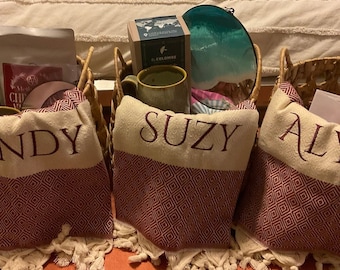May include: Three personalized decorative towels with names embroidered in burgundy thread. Each towel has a cream-colored top with a burgundy geometric pattern and fringe. Baskets hold a mug, coffee, and a teal-colored item.