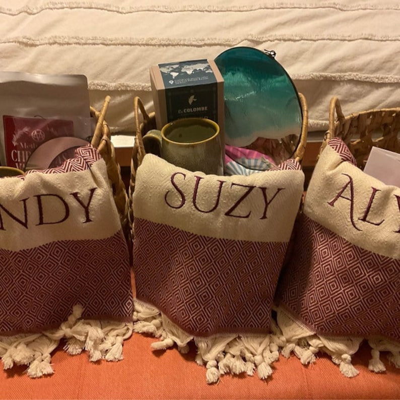 May include: Three wicker baskets with handles, each containing a gift. The baskets are lined with a cream and burgundy patterned fabric. The baskets are labelled "Andy", "Suzy", and "Ali" in burgundy embroidery.