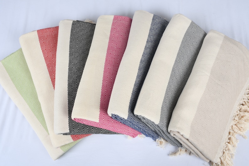 May include: A set of seven Turkish towels in various colors, including green, red, black, and blue. The towels are made of cotton and have a fringe on the ends. They are folded and arranged in a fan shape.