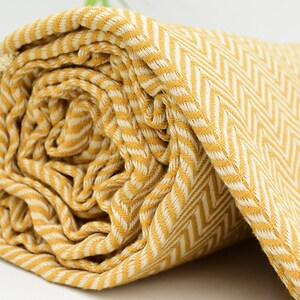 Op de afbeelding: Een gele en witte gestreepte deken met een herringbone-patroon. De deken is opgerold en de stof is zacht en textuur.
