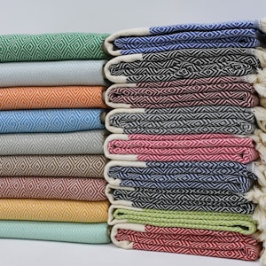 May include: A stack of colourful Turkish towels with diamond patterns and fringe. The towels are folded and arranged in a row, with the fringe facing outwards. The towels are in various colours, including green, blue, red, orange, and yellow.