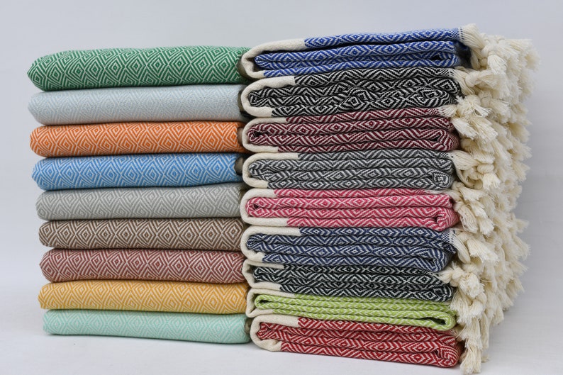 May include: A stack of colourful Turkish towels with diamond patterns and fringe. The towels are folded and arranged in a row, with the fringe facing outwards. The towels are in various colours, including green, blue, red, orange, and yellow.