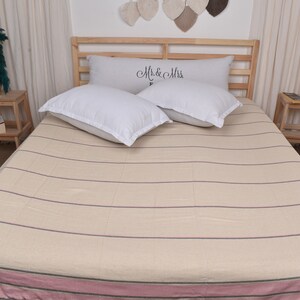 Puede incluir: Un cubrecama de color crema con finas rayas rosas y un borde rosa más grueso. Dos almohadas blancas con el texto "Mr. & Mrs." están en la cama.