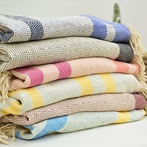 Op de afbeelding: Een stapel kleurrijke Turkse handdoeken met gefranjerde randen. De handdoeken hebben geometrische patronen in verschillende kleuren, waaronder blauw, roze, geel en rood. De handdoeken zijn gemaakt van een zacht, getextureerd materiaal.