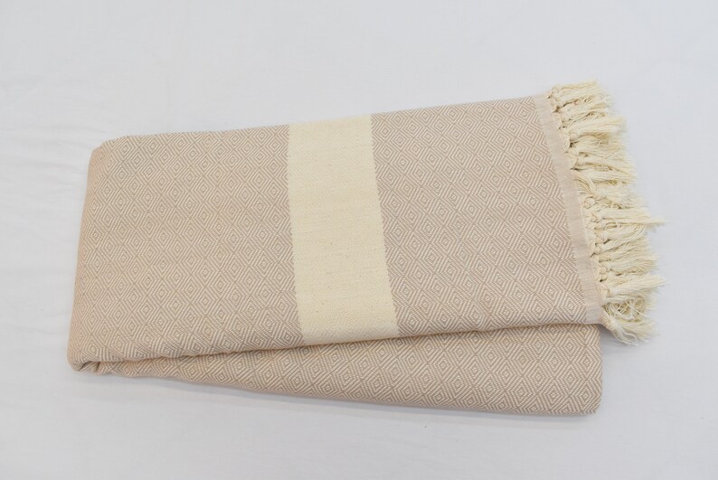 May include: A beige Turkish towel with a white stripe down the center and fringe on the edges.