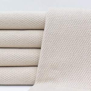 May include: A stack of four beige fabric towels with a textured weave. The towels are folded and stacked on a white surface.