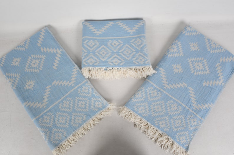 Pu&ograve; includere: Tre asciugamani di colore azzurro chiaro con un motivo geometrico bianco e frange. Gli asciugamani sono piegati e disposti a forma di triangolo.