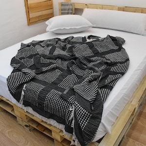 May include: A dark gray and white patterned blanket draped over a bed. The blanket has a textured design with fringe along the edge. The bed frame is made of wooden pallets, and the bedding is white.