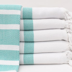 May include: A stack of six white towels with teal stripes and fringe edges. The towels are folded and stacked on a white surface.