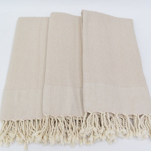 May include: Three beige Turkish towels with fringed edges. The towels are folded and arranged on a white surface.