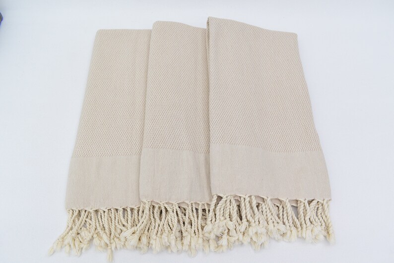 May include: Three beige Turkish towels with fringed edges. The towels are folded and arranged on a white surface.