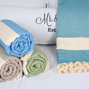 May include: Four rolled up Turkish towels in different colors, including blue, green, and beige, with white fringe. The towels are on a white surface with a white pillow with the text "Mr. & Mrs. Est." in black.