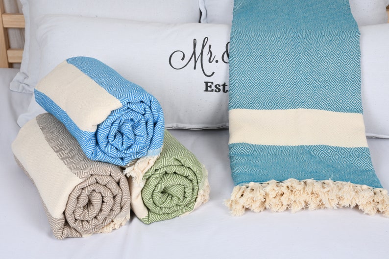 May include: Four rolled up Turkish towels in different colors, including blue, green, and beige, with white fringe. The towels are on a white surface with a white pillow with the text "Mr. & Mrs. Est." in black.