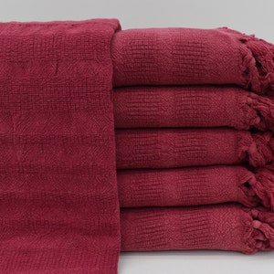 Puede incluir: Una pila de seis toallas de algodón burdeos con bordes con flecos. Las toallas están dobladas y apiladas sobre una superficie blanca.