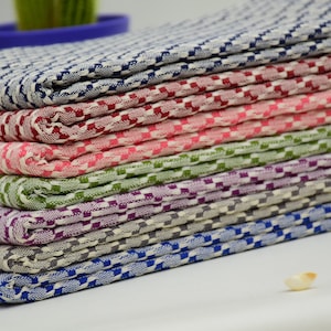 Op de afbeelding: Een stapel gevouwen stoffen in verschillende kleuren, waaronder blauw, rood, roze, groen, paars en grijs. De stof heeft een geruit patroon. Het materiaal lijkt katoen of linnen te zijn.