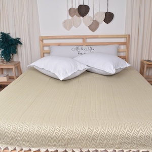Puede incluir: Una cama con estructura de madera clara, cubierta con una manta beige con estampado de espiga. Dos almohadas blancas descansan sobre la cama, con una almohada decorativa detrás que dice "Mr. & Mrs. Est. 2017". Dos mesitas de noche de madera a ambos lados de la cama.