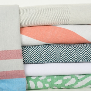 May include: A stack of six towels in various colors and patterns, including a white towel with a herringbone pattern, a coral pink towel with white stripes, a white towel with a green and white abstract pattern, and a gray towel with a red stripe.