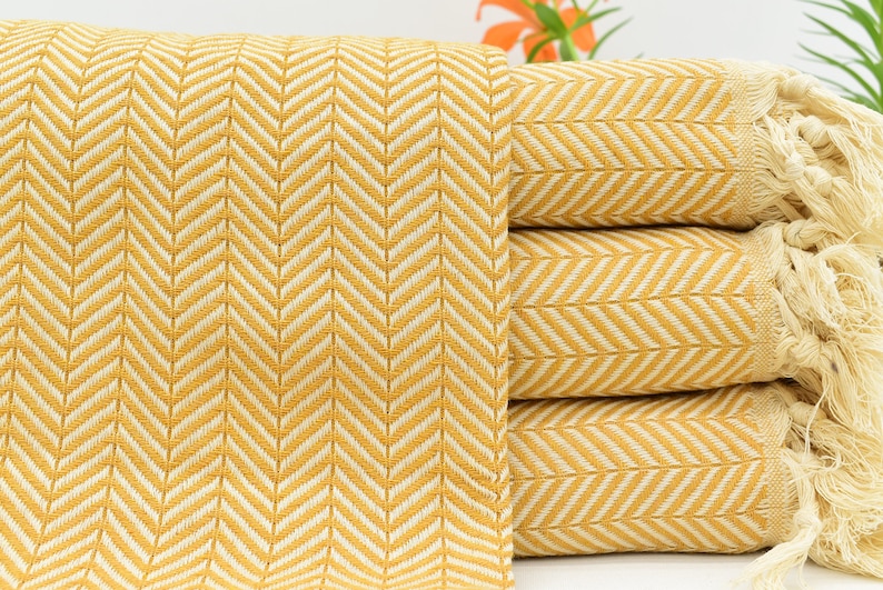 Op de afbeelding: Een stapel gele en witte gestreepte dekens met franjes aan de randen. De dekens zijn gemaakt van een zachte, geweven stof en hebben een herringbone-patroon.