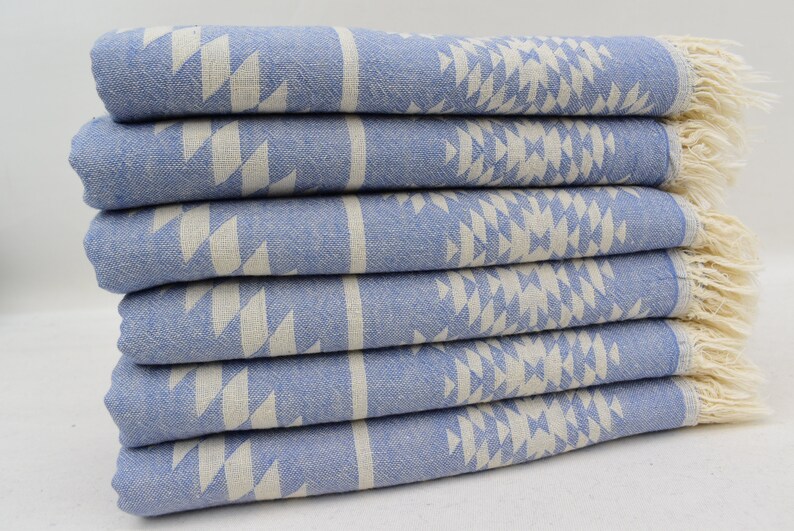 Peut inclure: Une pile de six serviettes de bain bleues et blanches &agrave; motifs avec des franges sur les bords. Les serviettes sont pli&eacute;es et empil&eacute;es sur une surface blanche.