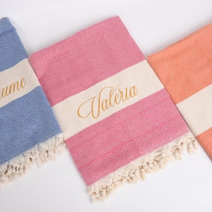May include: Three personalised beach towels with fringe. The towels are blue, pink and orange with a white stripe and gold embroidery. The names "Guillaume", "Valeria" and "Alva" are embroidered on the towels.