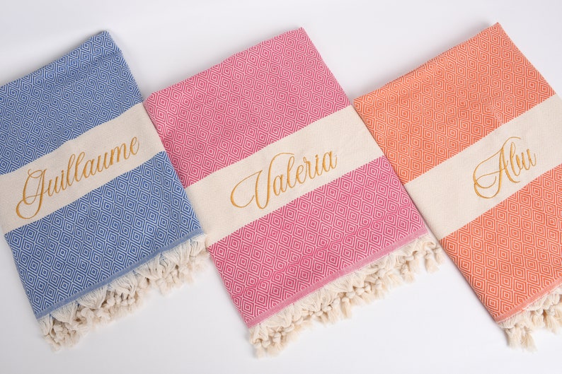 May include: Three personalised beach towels with fringe. The towels are blue, pink and orange with a white stripe and gold embroidery. The names "Guillaume", "Valeria" and "Alva" are embroidered on the towels.