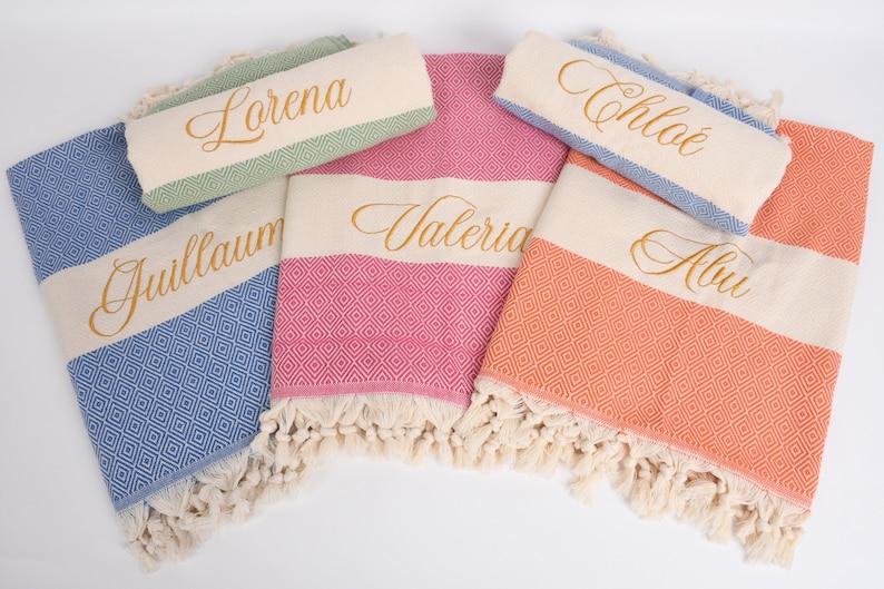 May include: Four personalised towels with different colours and patterns. The towels have the names "Lorena", "Guillaume", "Valeria", and "Alua" embroidered in gold lettering.