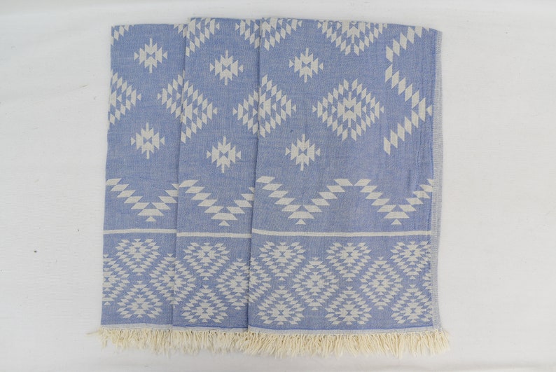 Peut inclure: Une serviette turque &agrave; motifs bleus et blancs avec une bordure en franges. La serviette pr&eacute;sente un motif g&eacute;om&eacute;trique avec des diamants et des triangles blancs sur un fond bleu clair.