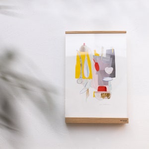 May include: Abstract art print in a wooden frame. The artwork features a colourful composition of yellow, red, grey, and white shapes. The print is displayed on a white wall, with a shadow from a plant in the foreground.