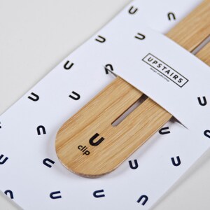 May include: A wooden clip with the word "clip" and the letter "U" printed on it. The clip is on a white background with a pattern of the letter "U" repeated. The text "UPSTAIRS" is printed on a white label.